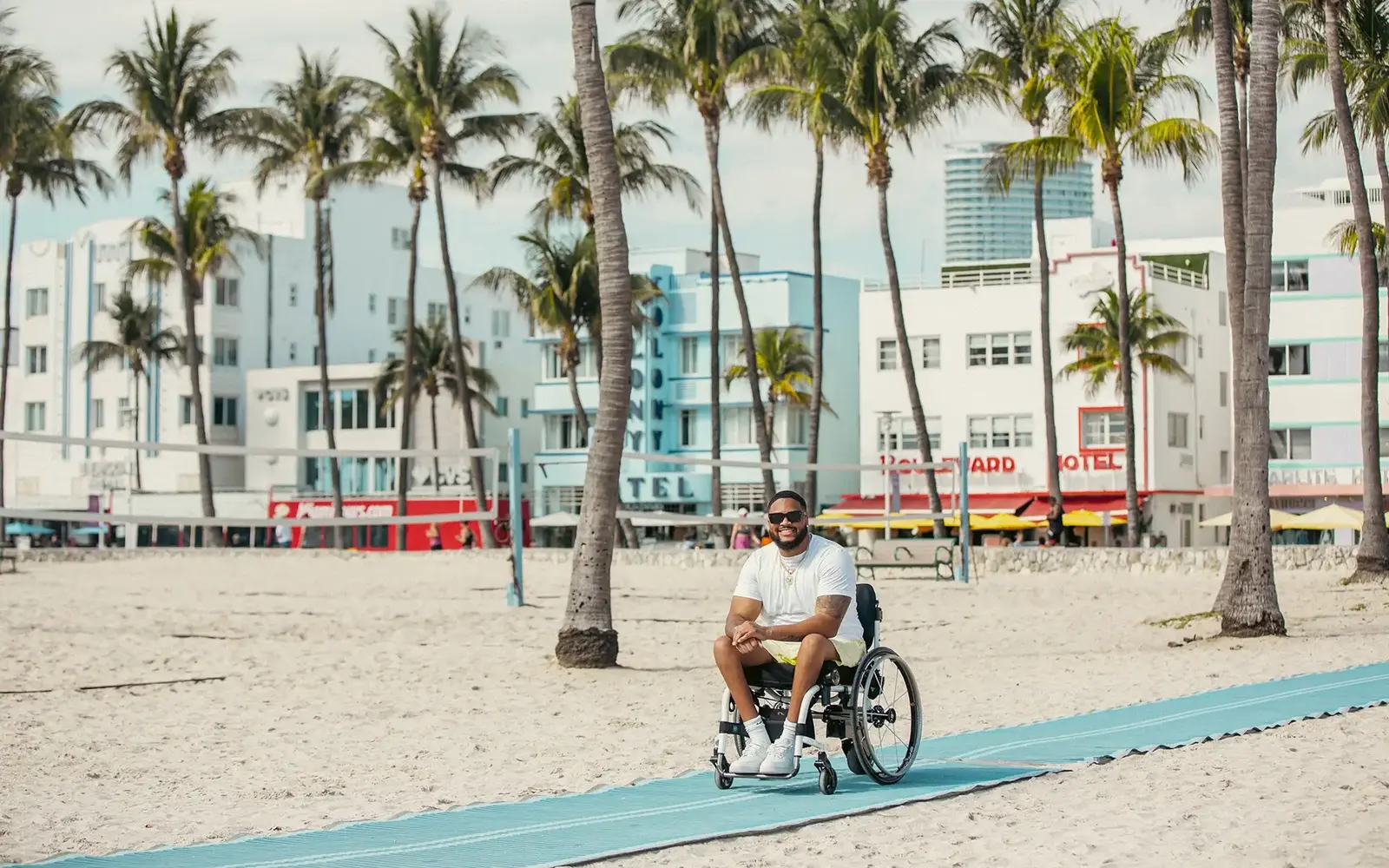 Man in a wheelchair on a beach mat with Art Deco hotels beyond
