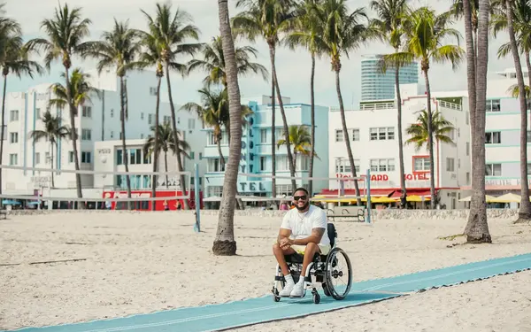 Man in a wheelchair on a beach mat with Art Deco hotels beyond