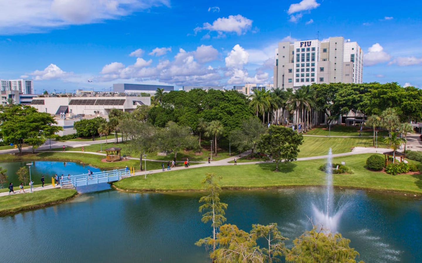 An aerial view of the FIU campus, showcasing lush greenery enveloping the buildings. A serene pond is crossed by a bridge, with students leisurely strolling to class An aerial view of the FIU campus, showcasing lush greenery enveloping the buildings. A serene pond is crossed by a bridge, with students leisurely strolling to class
